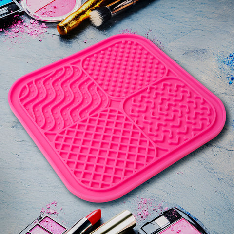 A rectangular pink silicone brush cleaning mat with various textured sections for different brush types, shown with makeup brushes and products around it.